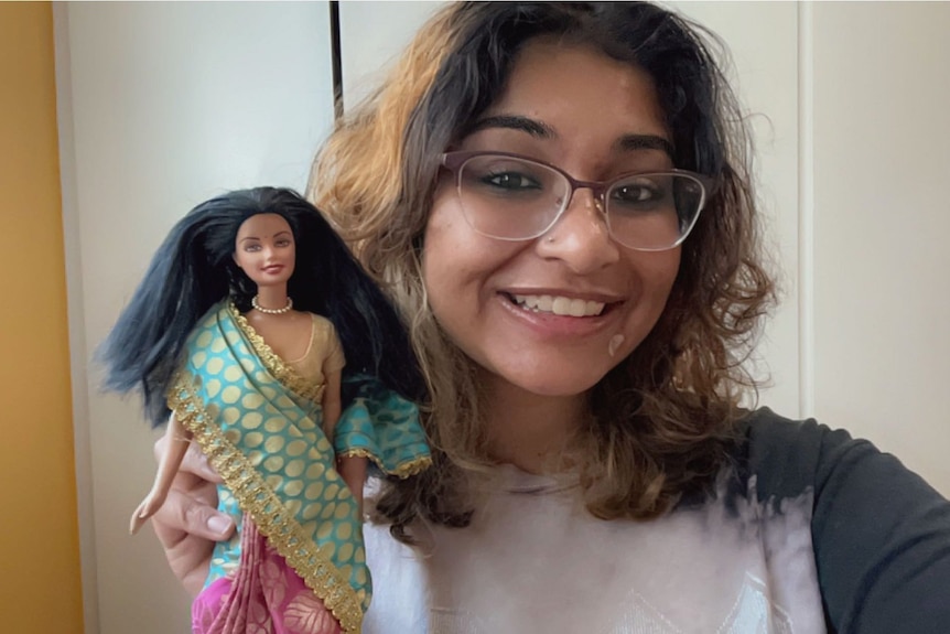 A woman smiling and holding up a Barbie doll with black hair, dressed in a saree
