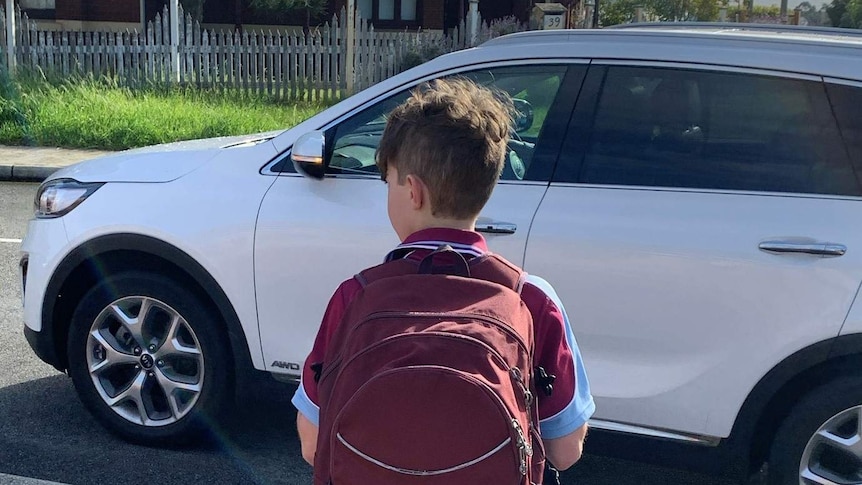 A schoolboy with a maroon backpack stands on the side of a road as a car passes