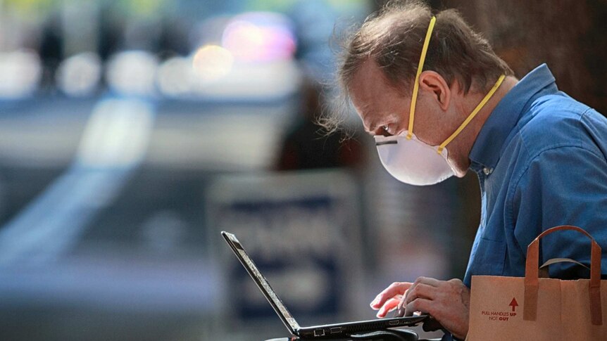A man sitting on a bench outside wears a face mask while working on a laptop.