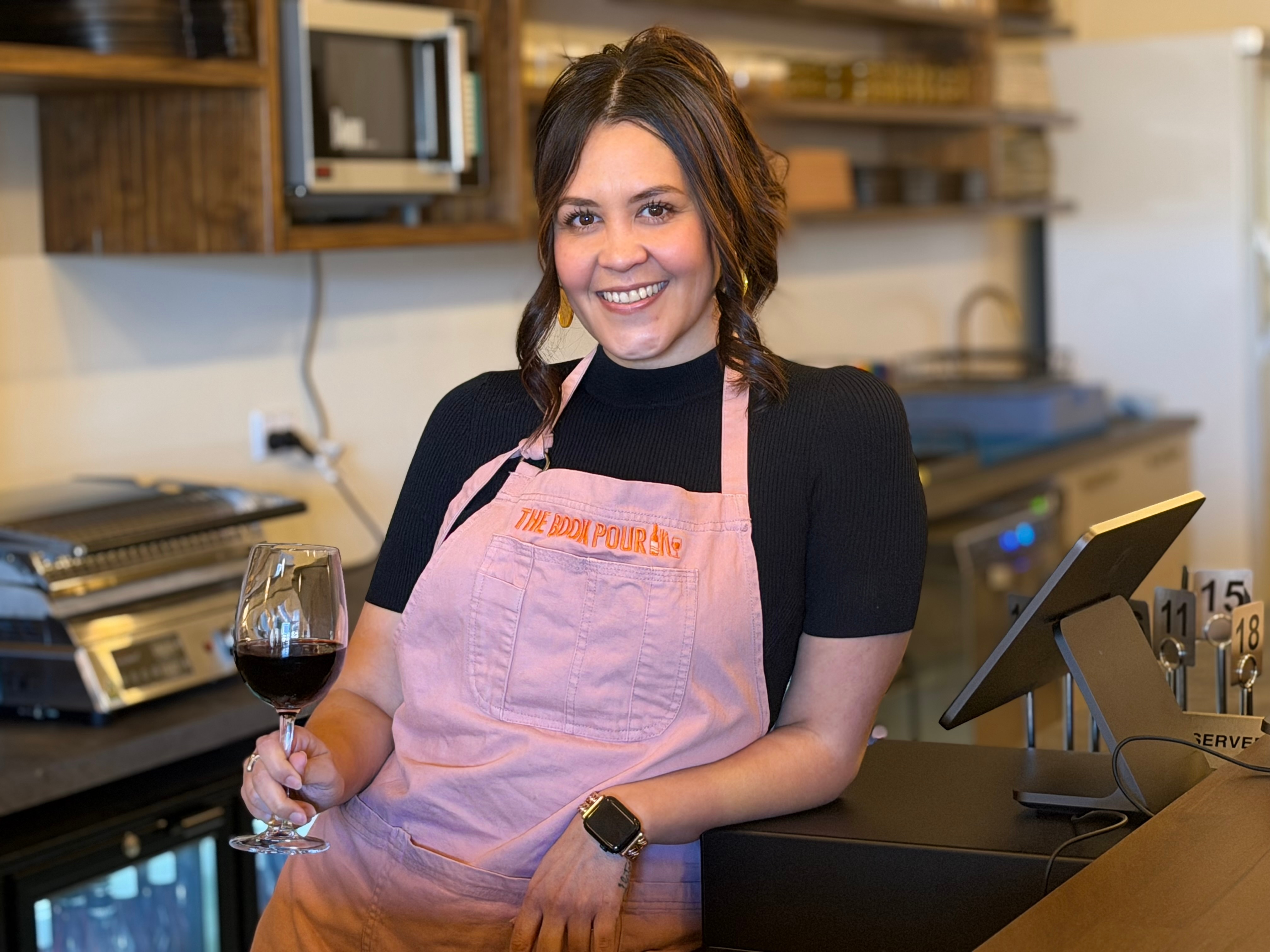 A woman leans on a bar near a cash register with a glass of wine in her hand.