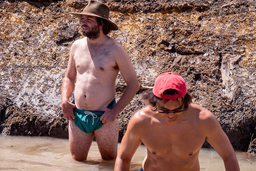 Two men shirtless in knee-deep muddy water.