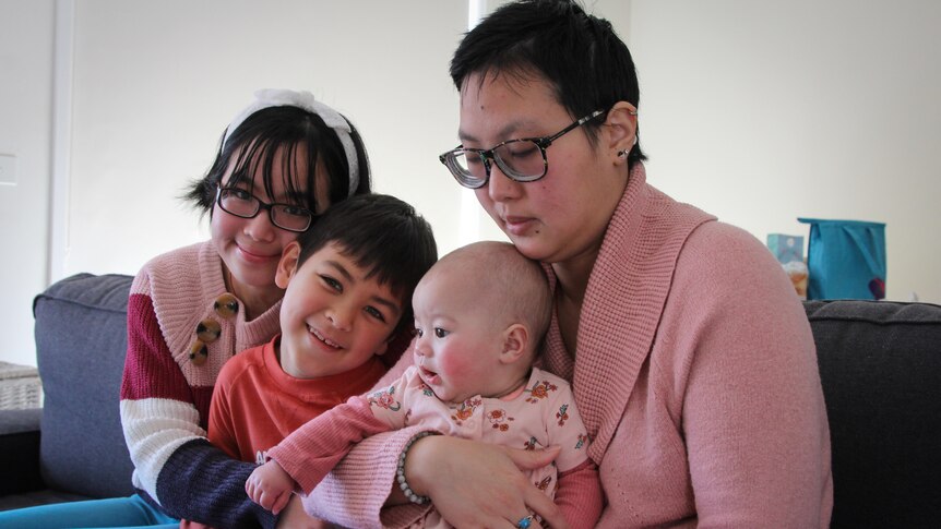 A woman with short black hair and glasses holds a baby girl, a 7 year old boy sits next to them and a young woman