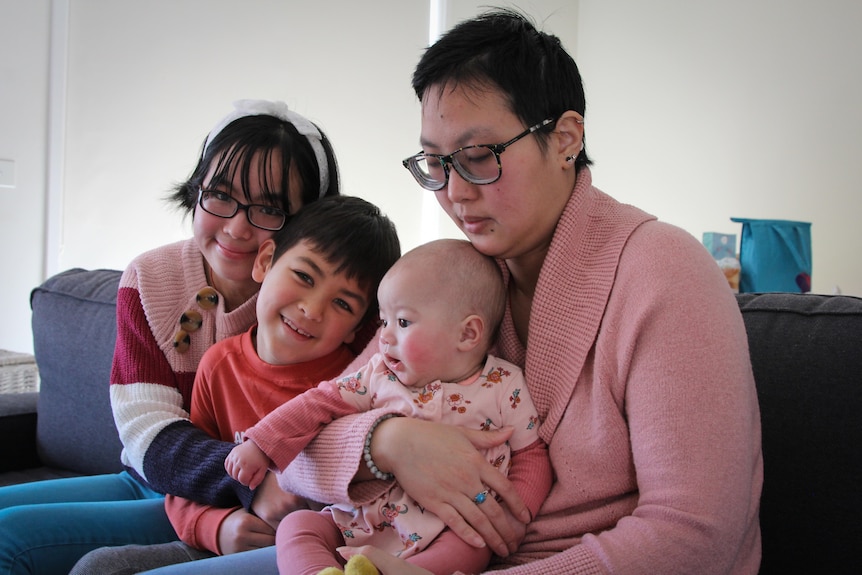 A woman with short black hair and glasses holds a baby girl, a 7 year old boy sits next to them and a young woman