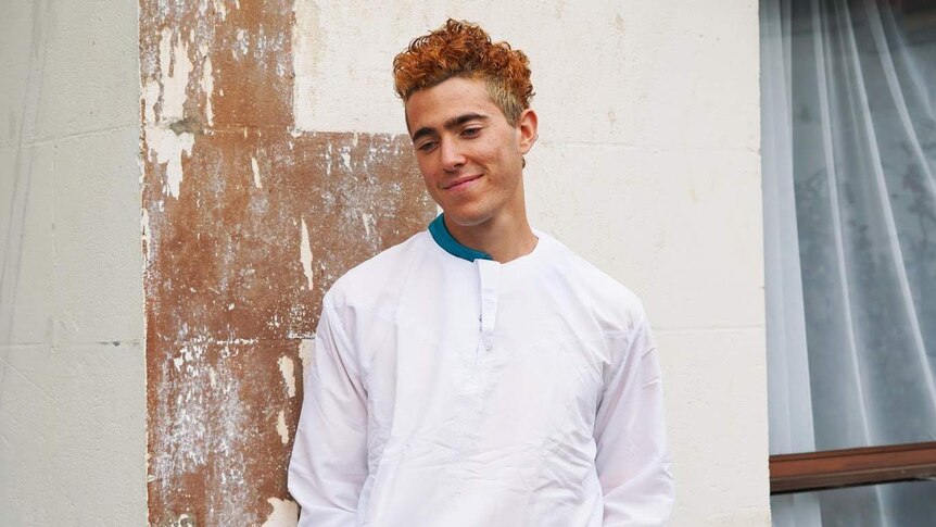 A young man with dyed orange hair stands smiling and looking at the ground wearing white long-sleeved clothing.