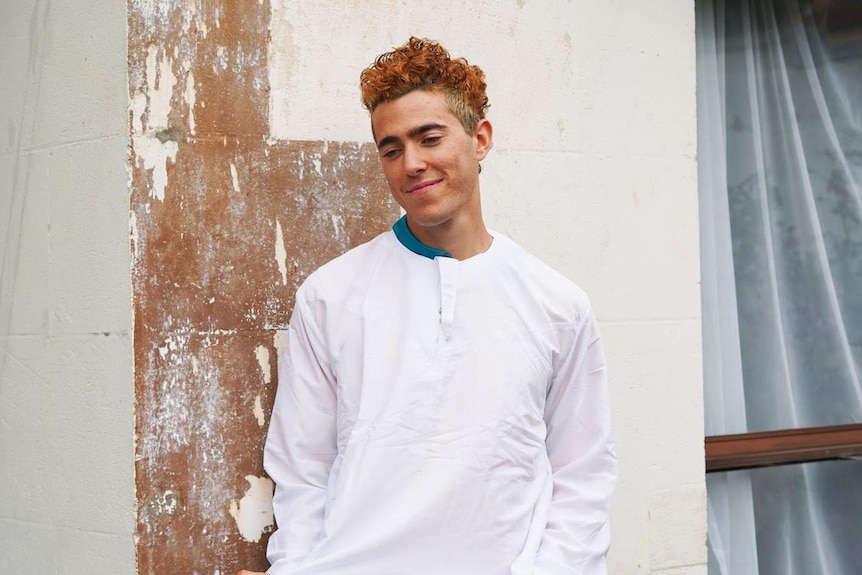 A young man with dyed orange hair stands smiling and looking at the ground wearing white long-sleeved clothing.