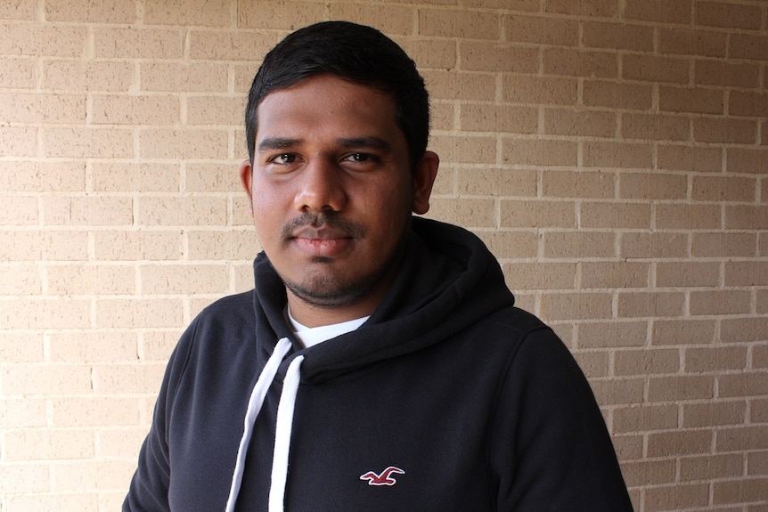 Heshan Perera wearing a black jumper and stands in front of a brown brick wall