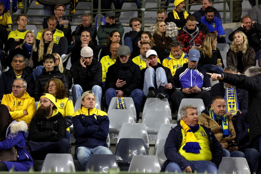 A crowd of Swedish football fans in the stands of a stadium, many on their phones