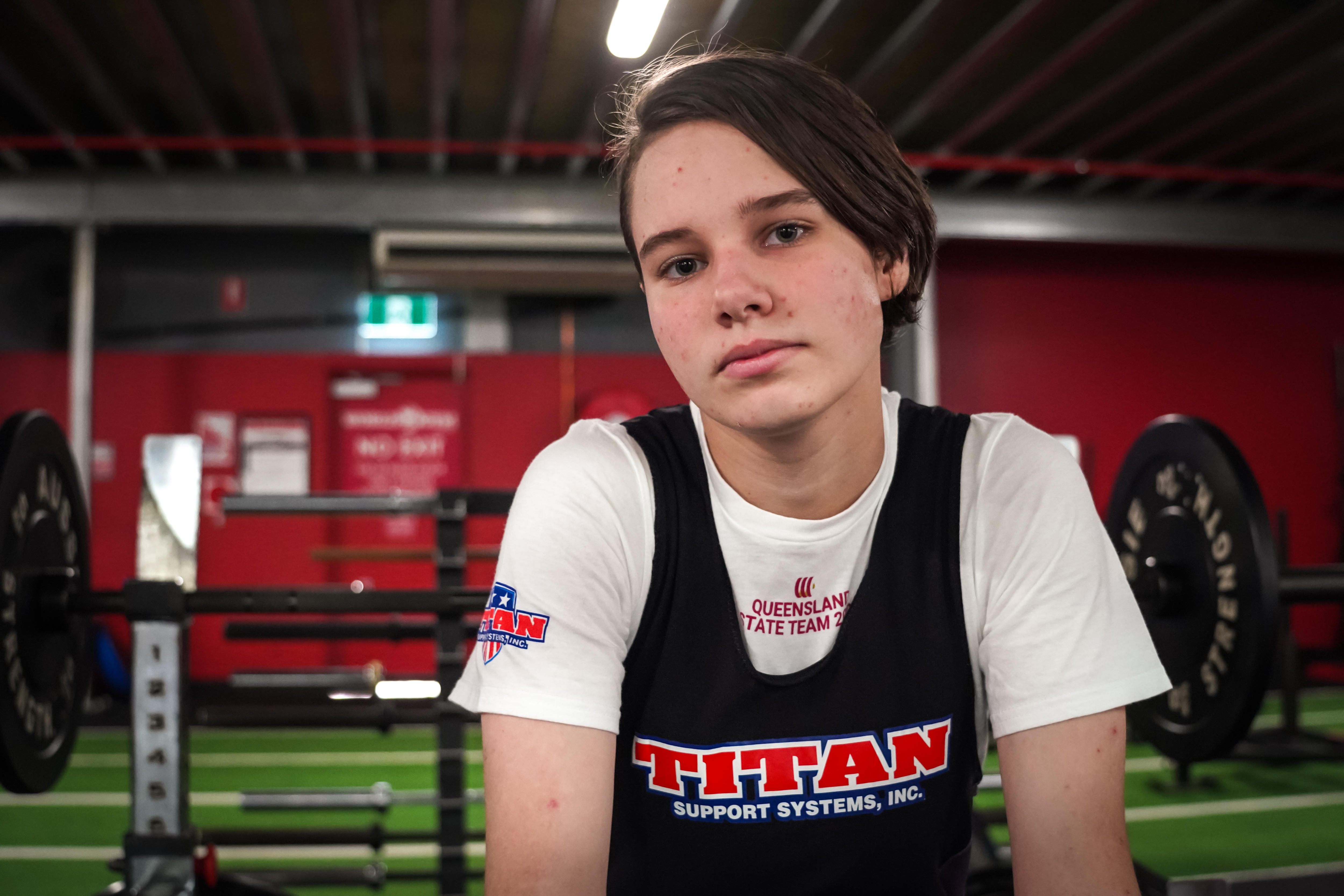 A teenage boy sits in a gym with weights and powerlifting equipment behind him.