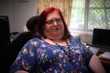 A woman with red hair poses for a photo while sitting in a wheelchair with a desk behind her in a room in a house.
