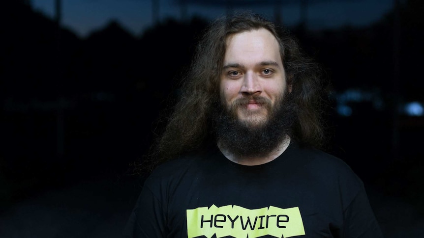 Portrait of a young man with long hair and a beard wearing a Heywire t-shirt.