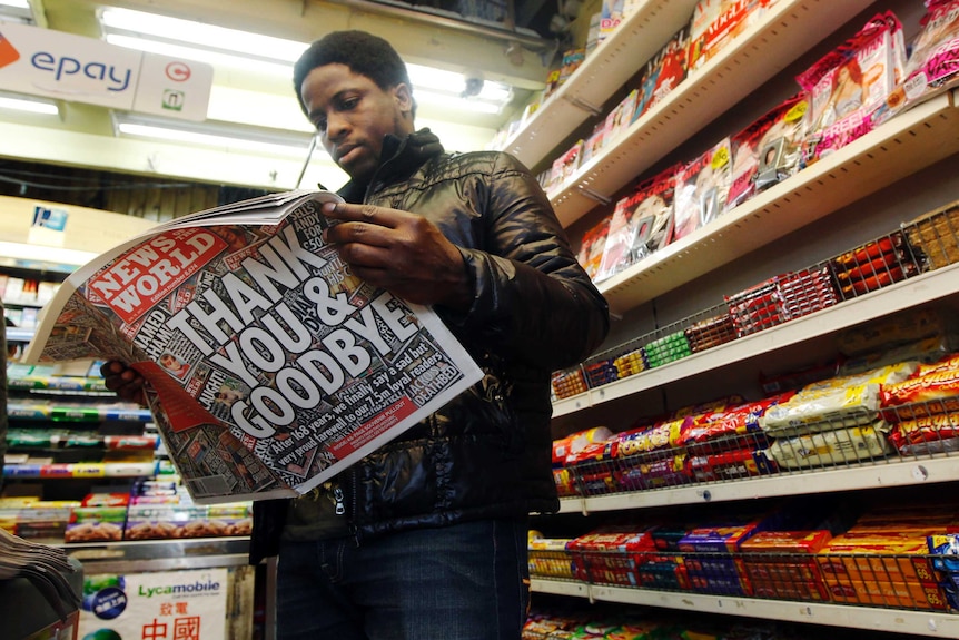 A man stands in a newsagents reading the News of the World newspaper in front of store shelves