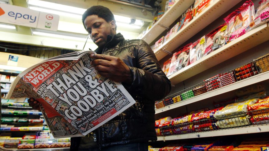 A man stands in a newsagents reading the News of the World newspaper in front of store shelves