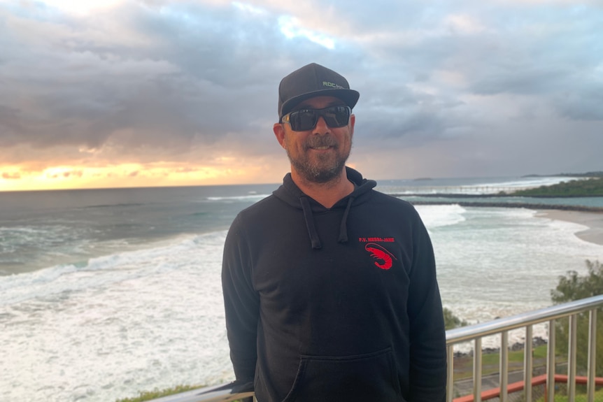 A man wearing a black cap and black hoodie with a logo of a red prawn standing in front of the ocean at sunrise