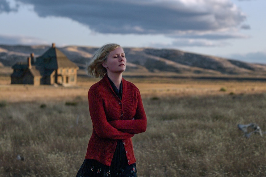 A 30-something woman in a red cardigan stands with her arms folded in a field, her eyes closed and blonde hair windswept