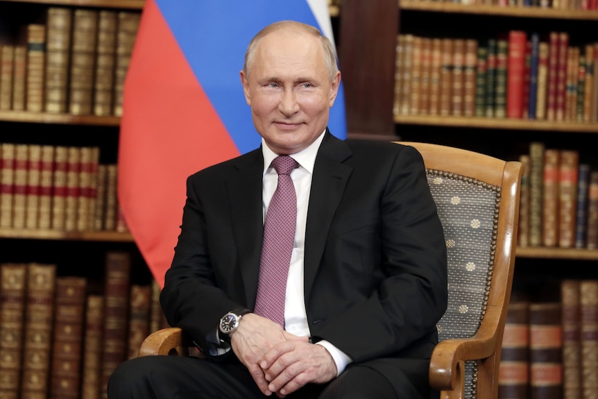 Vladimir Putin gives a slight smile in front of a Russian flag and a bookcase