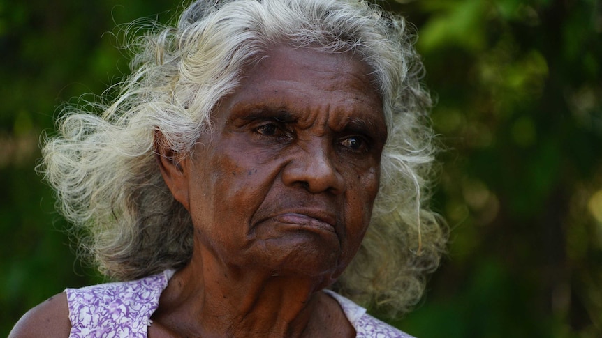 A medium close-up of an elderly Aboriginal woman, who is standing talking to a reporter but looking off camera.