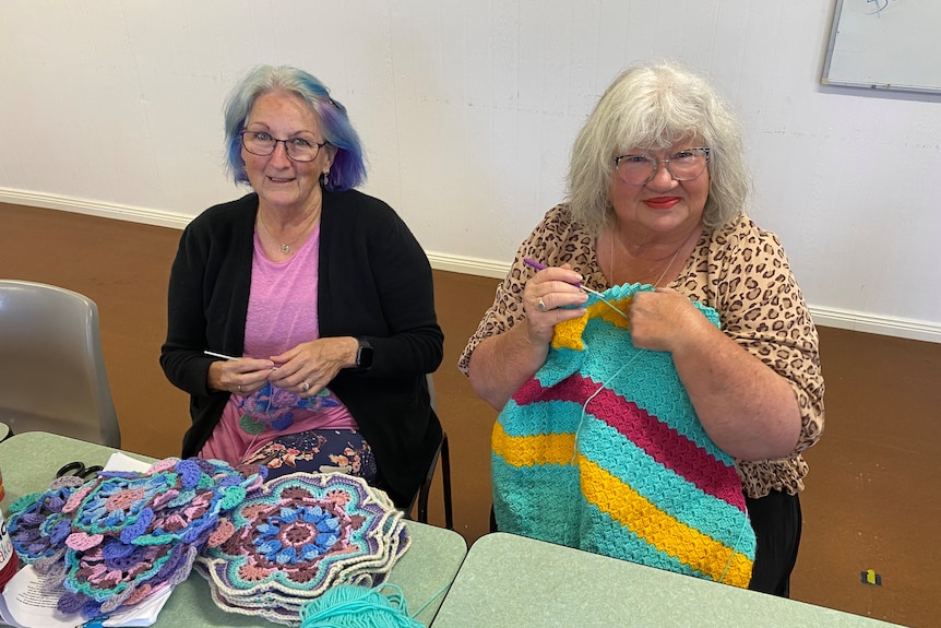 Two women sit side by side, smiling as they hold crochet projects