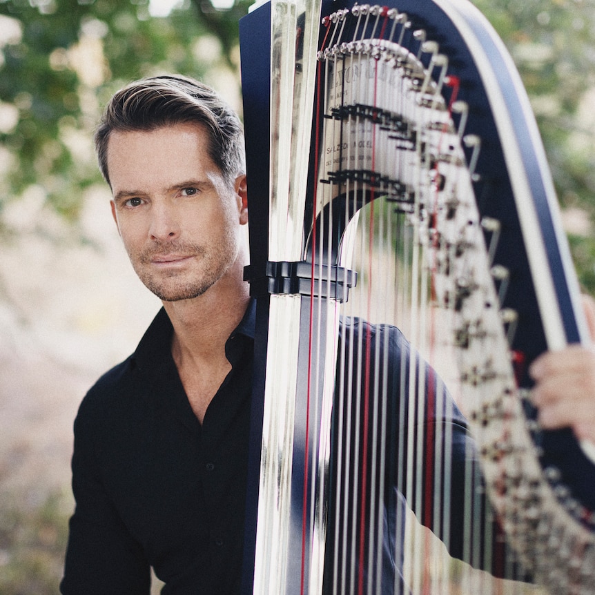A man in a black shirt stands with his arm around a blue concert harp against a background of trees.