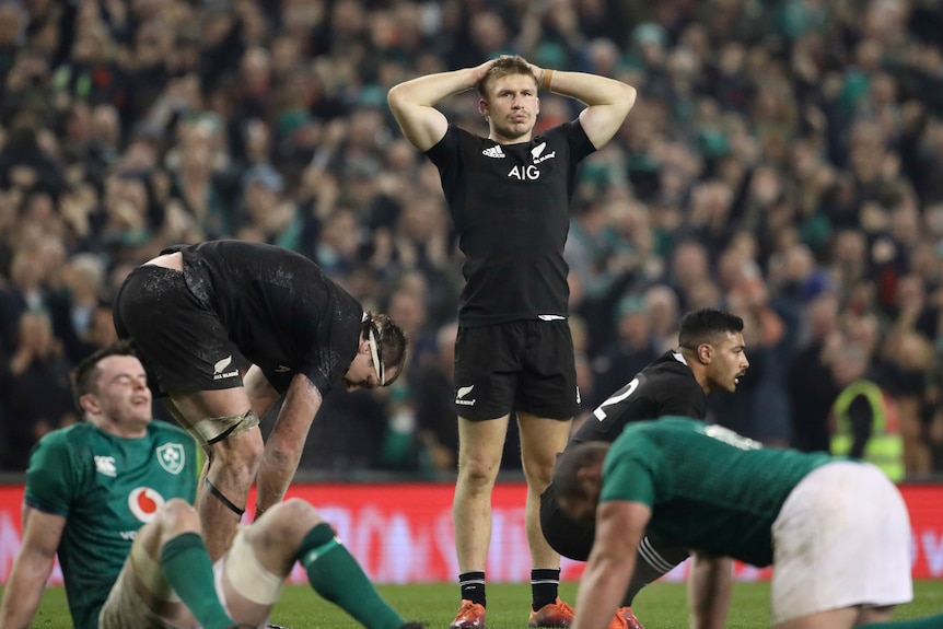 A man in a black rugby jersey stands with his hands on his head with other men on their haunches around him