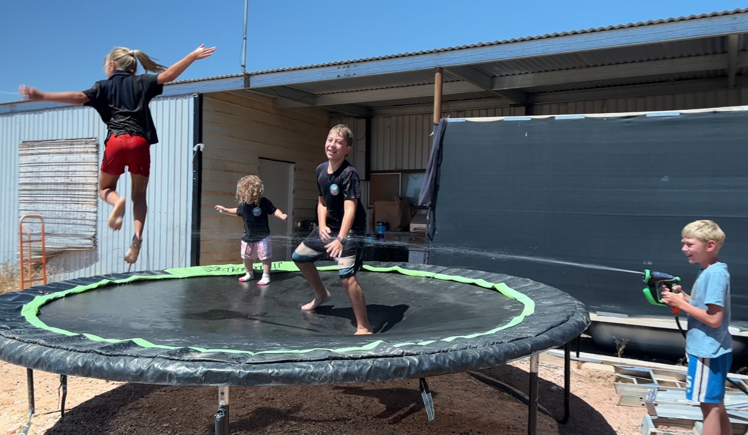 Four kids of varying ages play on a trampoline under a blue sky.