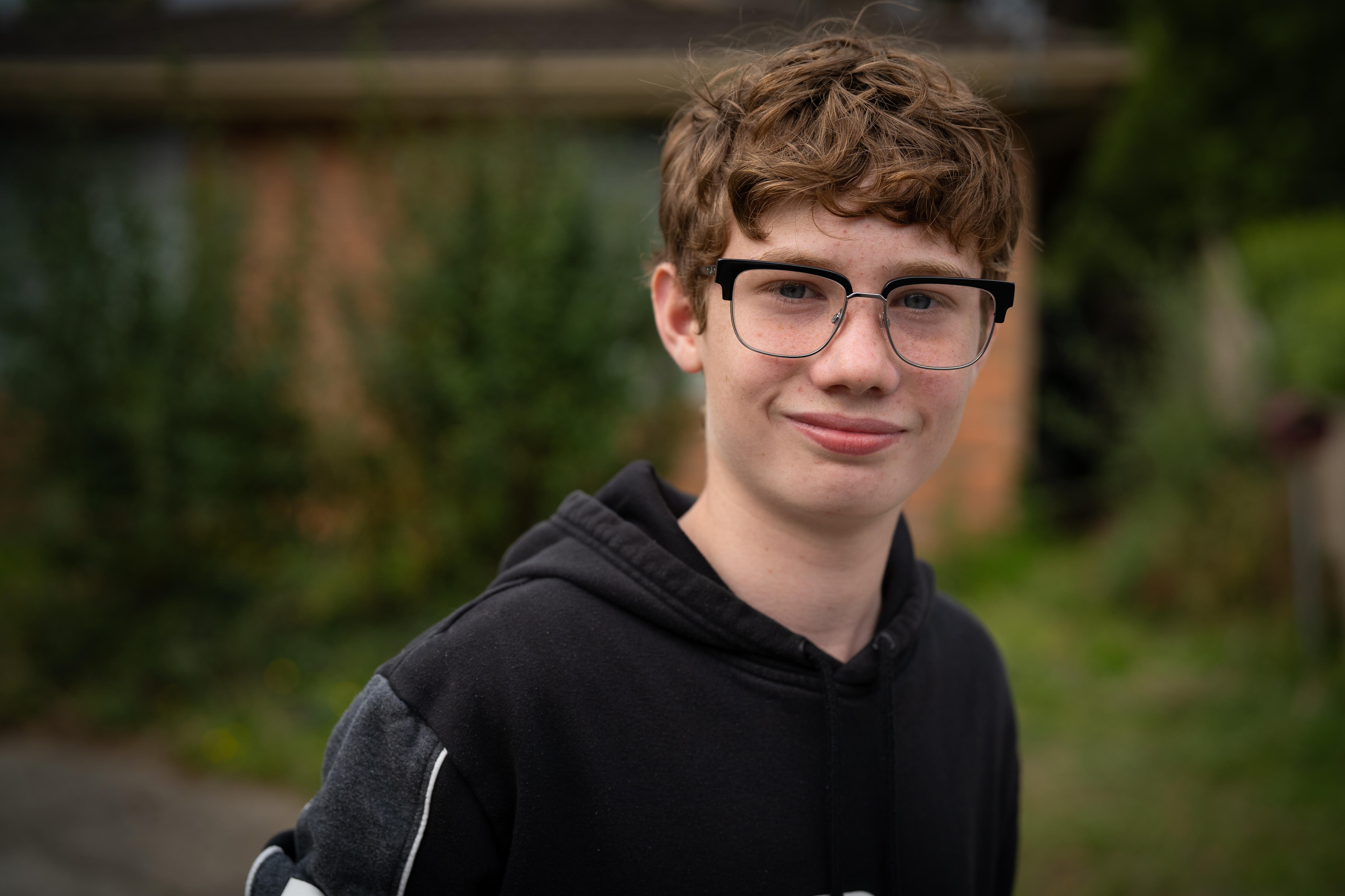 A red‑haired teenage boy wearing glasses smiles while standing in front of a house, dressed in a black jumper.