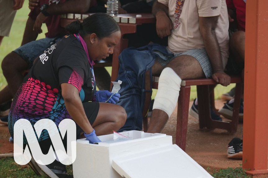 A woman crouches down near a crate of medical supplies next to a man's bandaged knee.