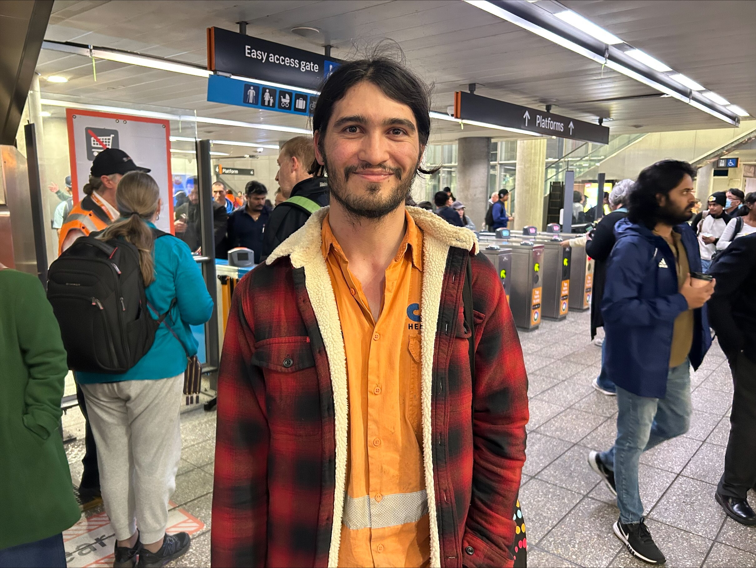 Un hombre joven con una frannelette y una camisa naranja sonríe de pie dentro de una estación de tren.