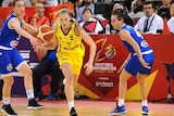 A blonde girl wearing a yellow uniform dribbles a basketball past two girls wearing blue