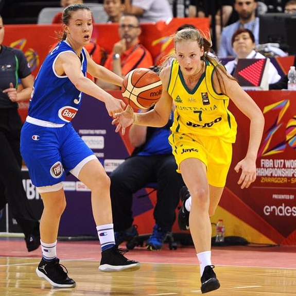 A blonde girl wearing a yellow uniform dribbles a basketball past two girls wearing blue