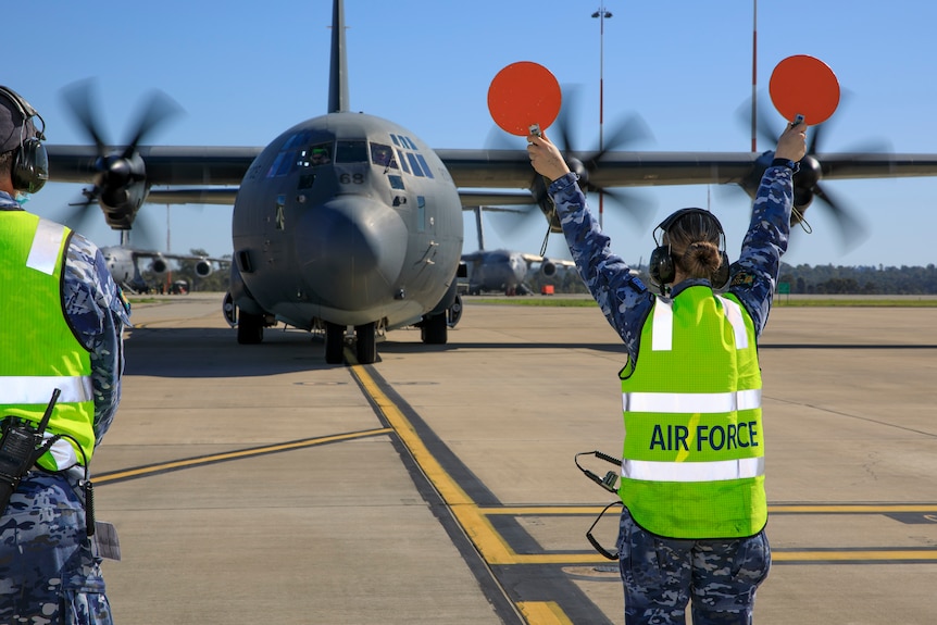 A large air force plane taxis on a runway in the background with an air force member directing it in the foreground