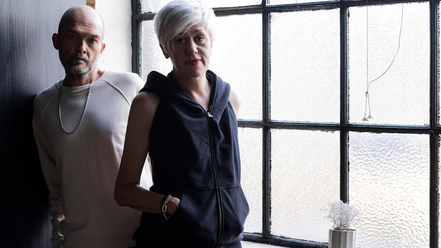 A bald man and silver haired woman stand in a dark room looking at the camera without expression