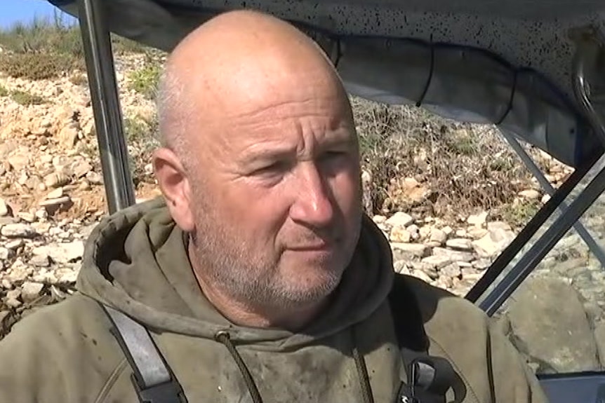 A man with a bald head wearing a dark green jumper stands next to his boat.