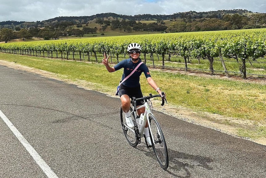 A girl on a bike putting up a peace sign with vineyards behind her.