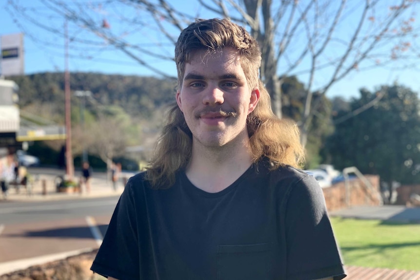 A young man in a black t-shirt smiling at the camera with a long mullet around his shoulders