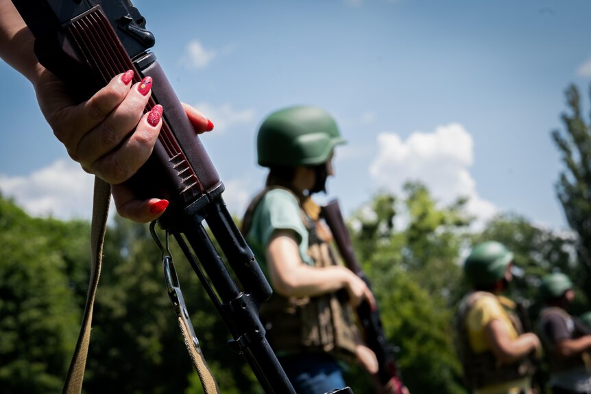 A woman's hand, fingernails painted in sparkly red, grips an assault rifle. Behind, people in helmets do the same
