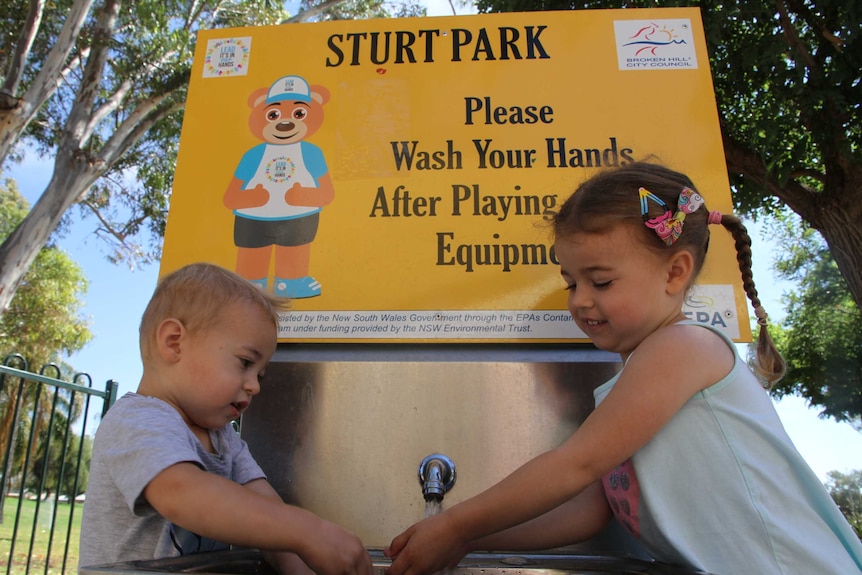 A young boy and girl stand on either side of the silver basin. Above their head reads a sign with a picture of lead ted