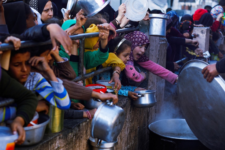 Children poke their heads and arms through a fence holding saucepans in front of a pot with steam rising from it.