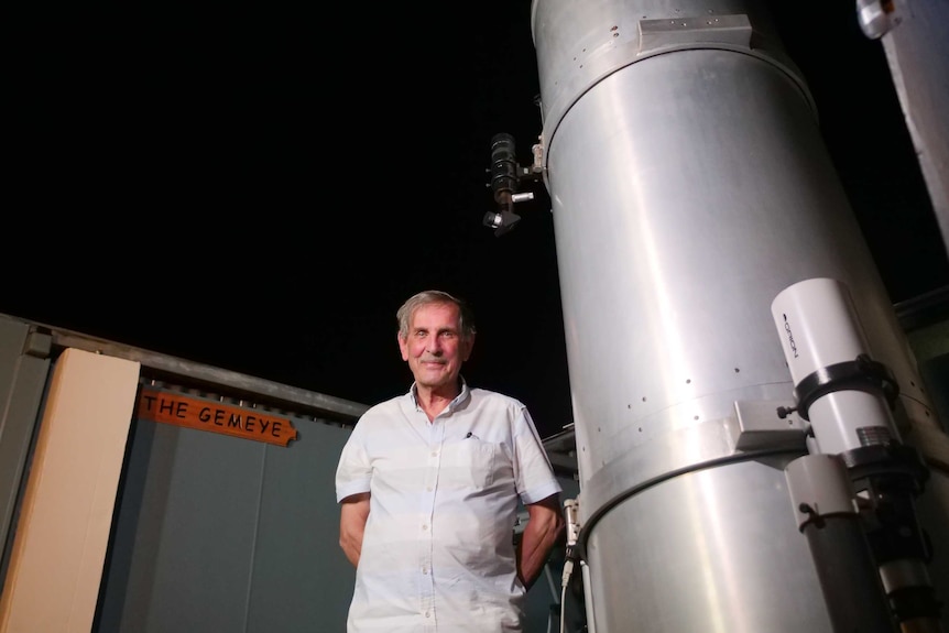 A man with grey hair stands beside a telescope that towers above him.