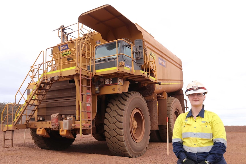 Woman in foreground front left wearing high vis gear including helmet and glasses with big yellow water truck in background