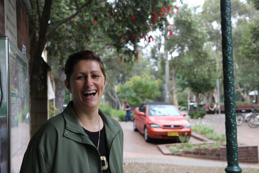 A woman laughs in front of a wattle tree and a parked car in the background.