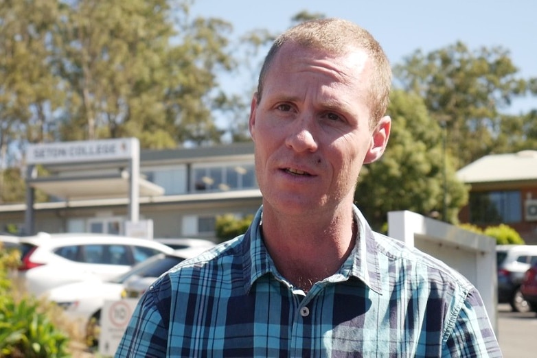 A man in a plaid shirt stands in front of a school.