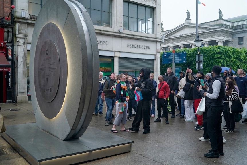 The Portal is pictured from behind as a crowd of people stands in front of it filming and seemingly laughing. 