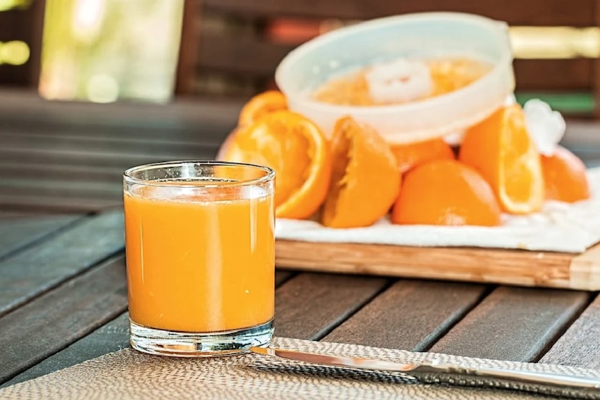 A glass of pulpy orange juice on a table in front of a pile of oranges and a hand-operated citrus juicer.