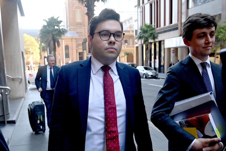 A man in a dark blue suit, red tie and glasses walks on a city street.