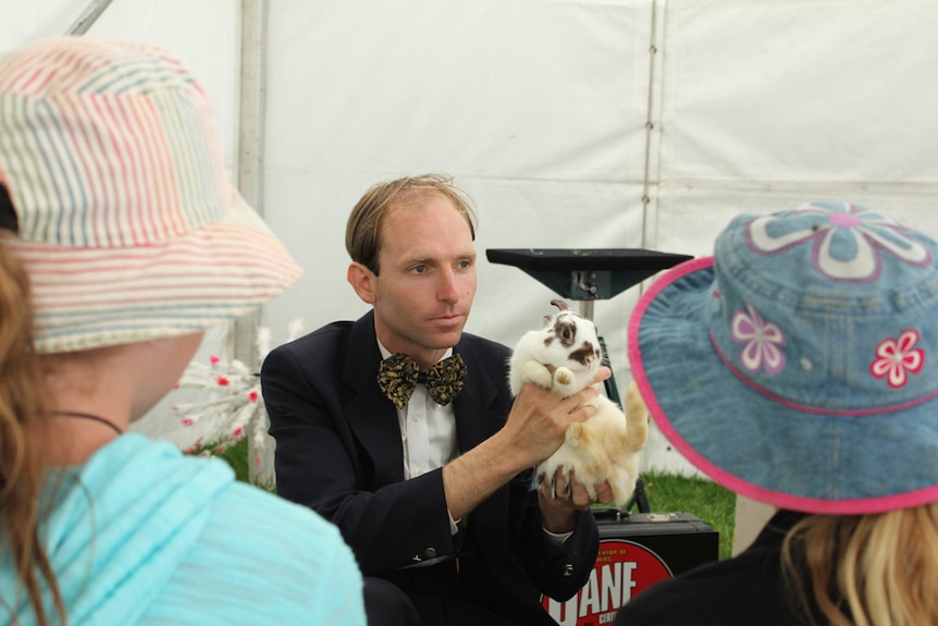 A man in a suit with a yellow and black bowtie holding a rabbit in front of a crowd.
