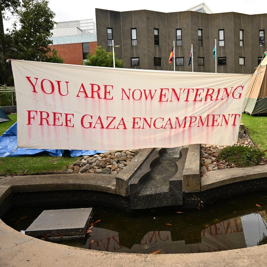 A large sign that says 'you are now entering free gaza encampment' with a few tents in the background and a Palestine flag.