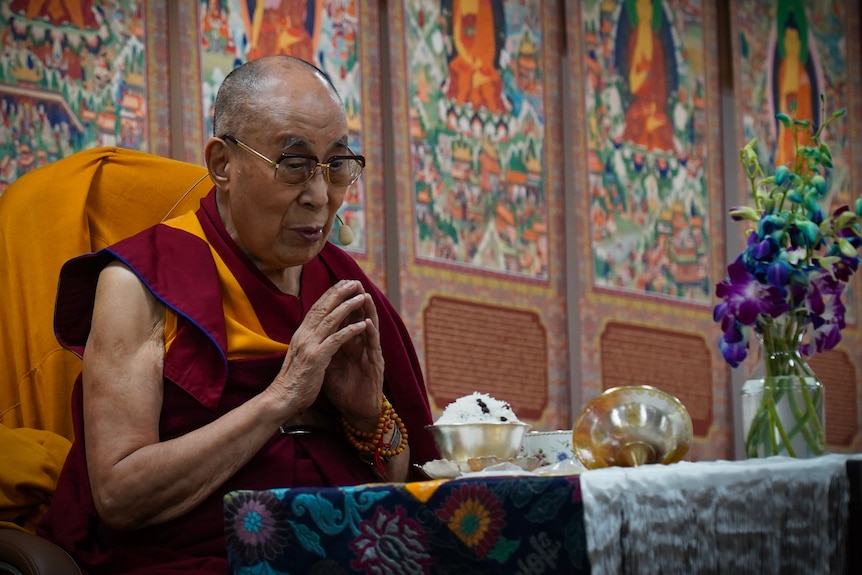 The Dalai Lama in his trademark orange and maroon robes puts his hands together in prayer at a table in front of colourful walls