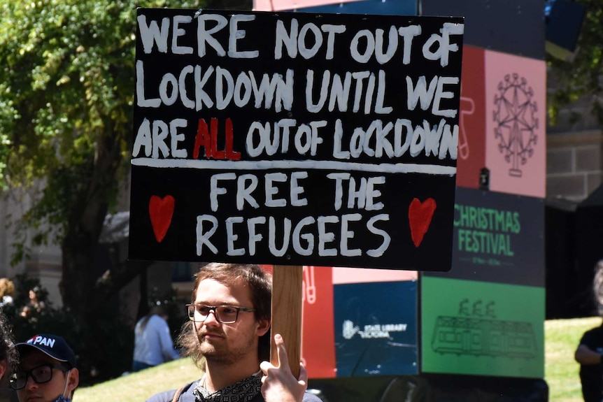 Two protesters with one holding a sign that says 'we're not out of lockdown until we are all out of lockdown'.