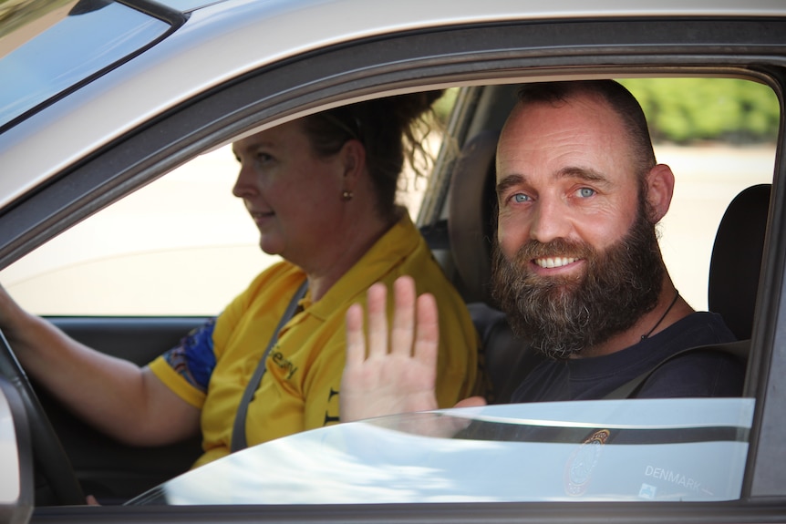 Thor sur le siège passager d'une voiture, sans son chapeau, sourit et salue, une femme en t-shirt jaune et bleu conduit.