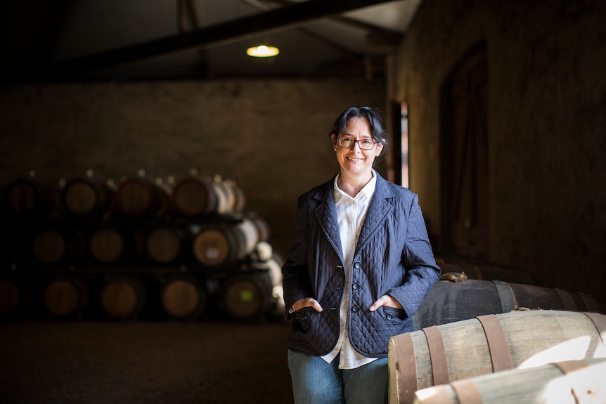 A white woman with brown hair and glasses, Louisa Rose, wears a navy blazer and smiles next to an oak barrel in a wine cellar.